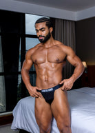 Man wearing a black 'Santo Ford' underwear set in a bedroom.